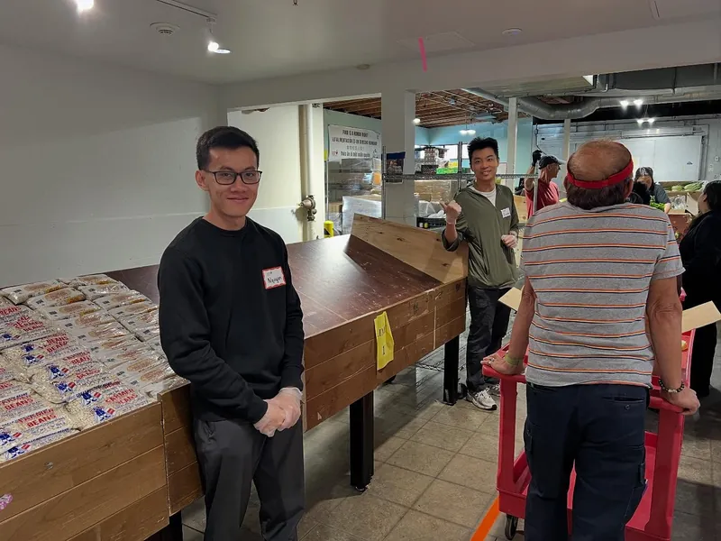 John volunteering at a food pantry, wearing a name tag and gloves beside a bin of dried beans
