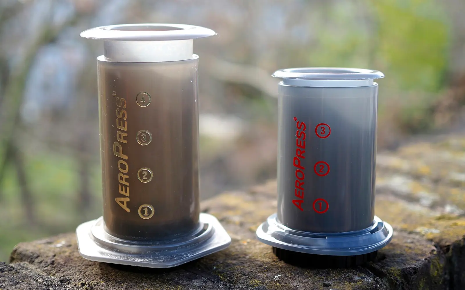 A 2015 AeroPress next to a 2020 AeroPress Go on a stone ledge outdoors
