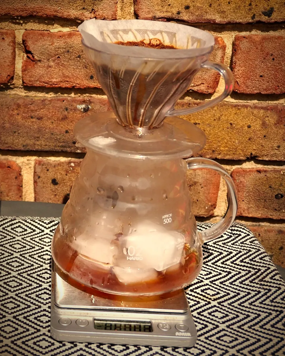 A V60 dripper on a glass server filled with ice, sitting on a digital scale in warm sunlight