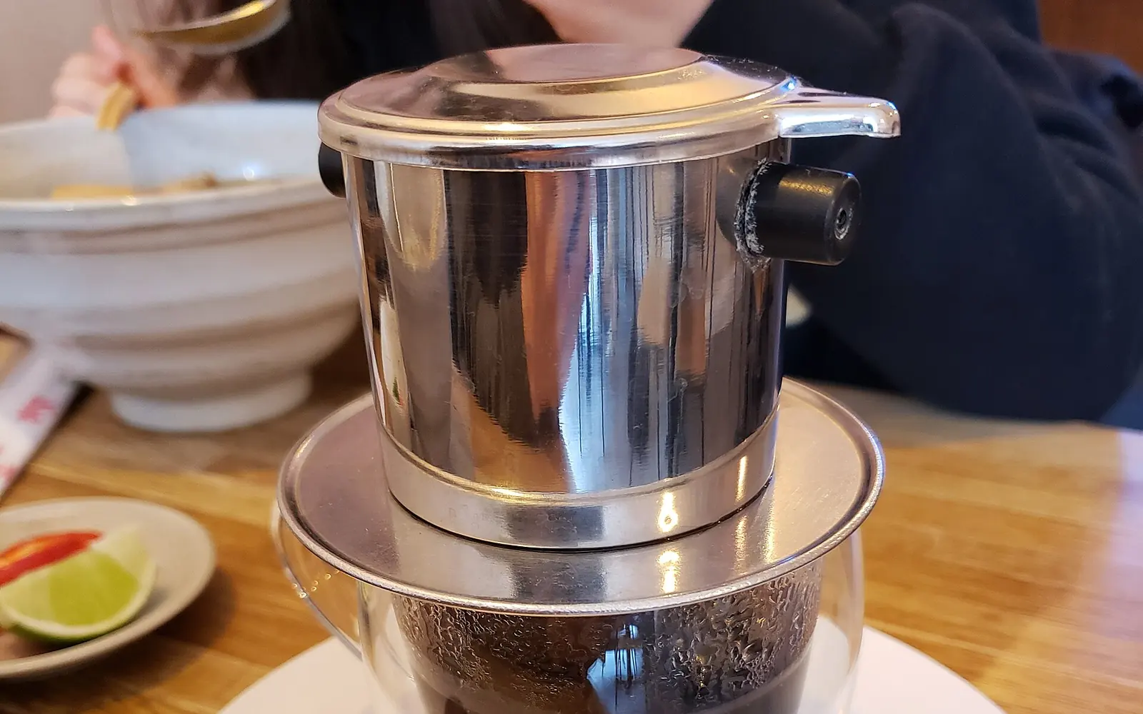 A stainless steel Vietnamese phin filter dripping coffee into a glass on a saucer