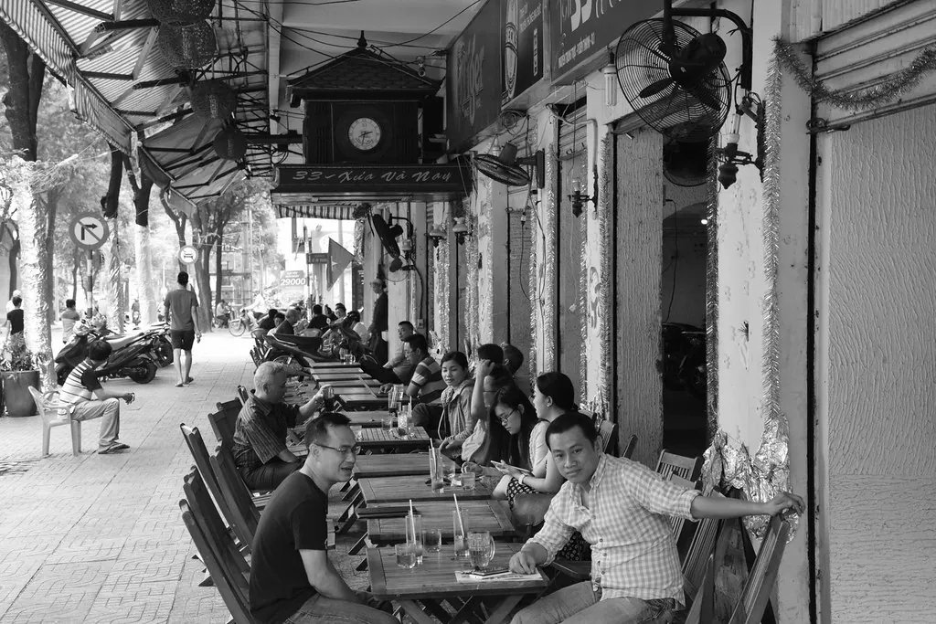 People sitting and talking at the long row of sidewalk tables of the Xưa và Nay café in Ho Chi Minh City, in black and white