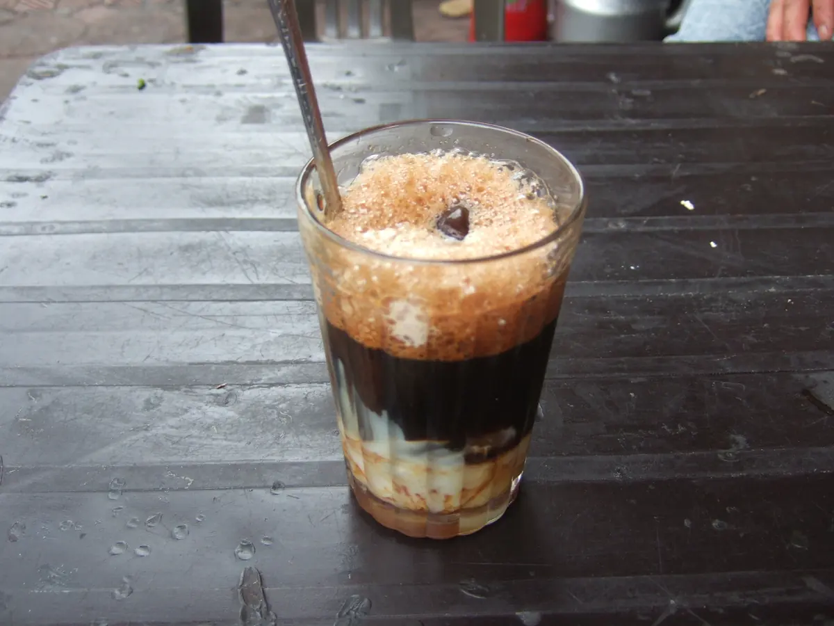A glass of cà phê sữa đá with layers of condensed milk, coffee, and ice, topped with foam