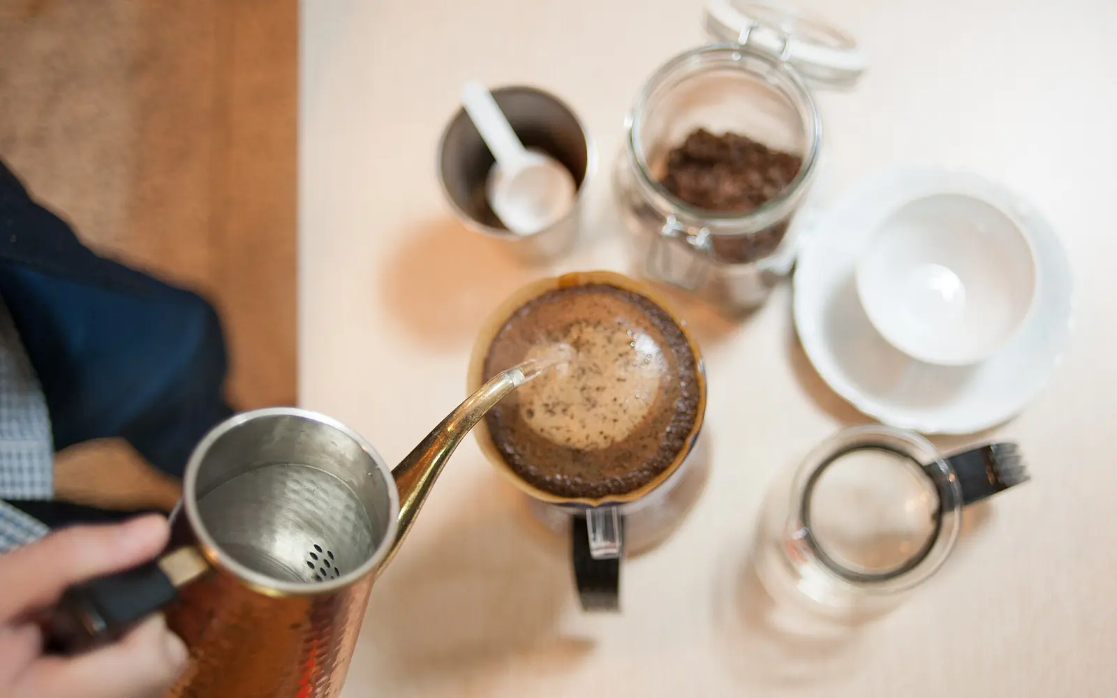 Hot water poured from a gooseneck kettle into a pour-over dripper, with a jar of beans and cups on a white table
