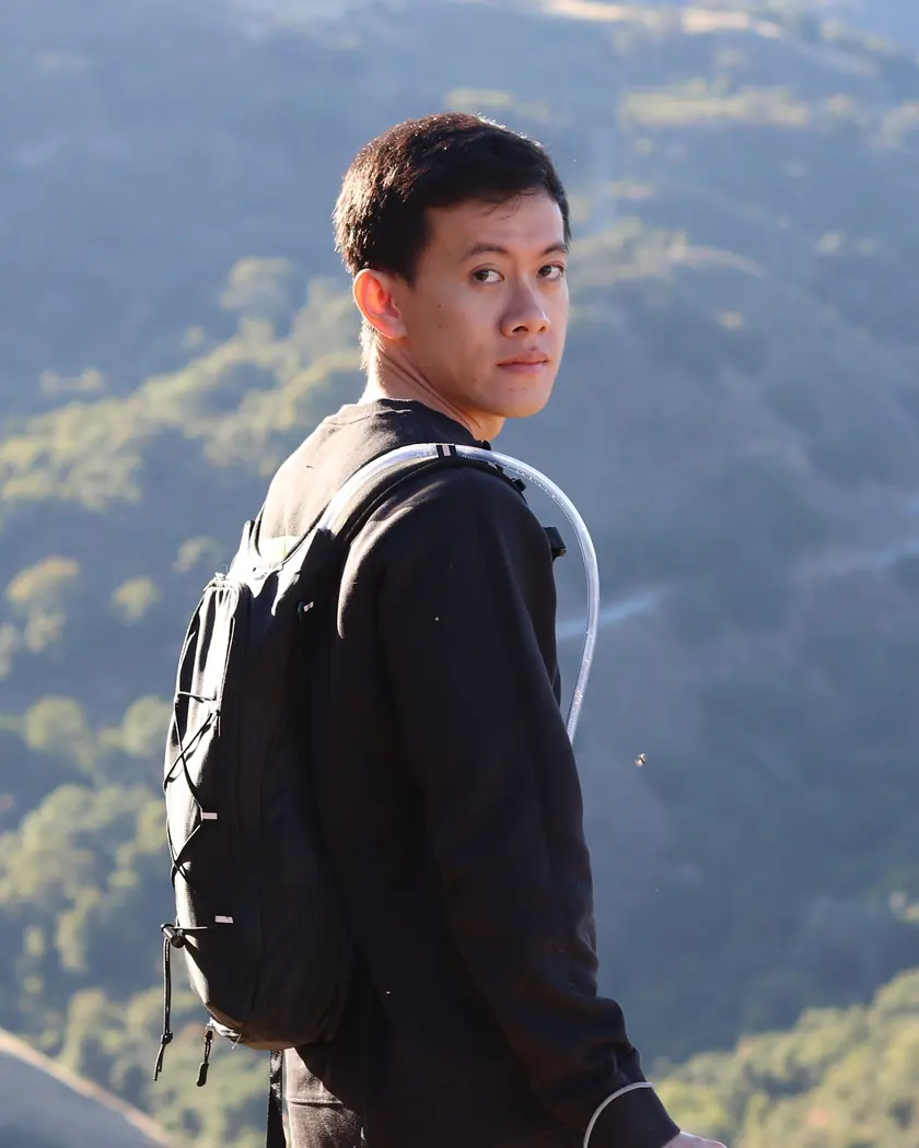 Portrait of John on a hiking trail, holding a camera, with rolling green hills behind him